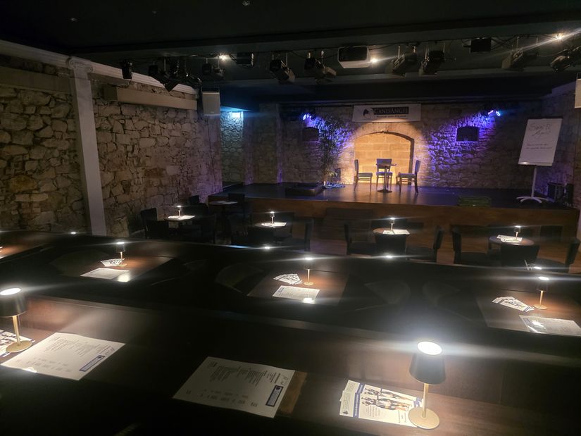 Blick auf die beleuchtete Bühne über die Zuschauerreihen hinweg, die aus Tischen und Stühlen bestehen; auf den Tischen kleine leuchtende Lampen