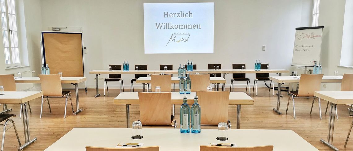 Eingedeckter Tagungsraum mit Tischen, die in kleinen Reihen hintereinander stehen, Blick auf eine Leinwand mit der Aufschrift „Herzlich willkommen“ und dem Logo des Halben Mondes
