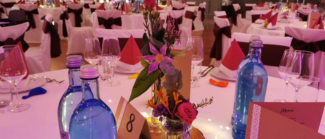 Helle festlich gedeckte Tische mit verzierten Menükarten, bunten Blumengestecken und Wasserflaschen aus blauem Glas; Stühle mit dunklen Schleifen über den weißen Hussen